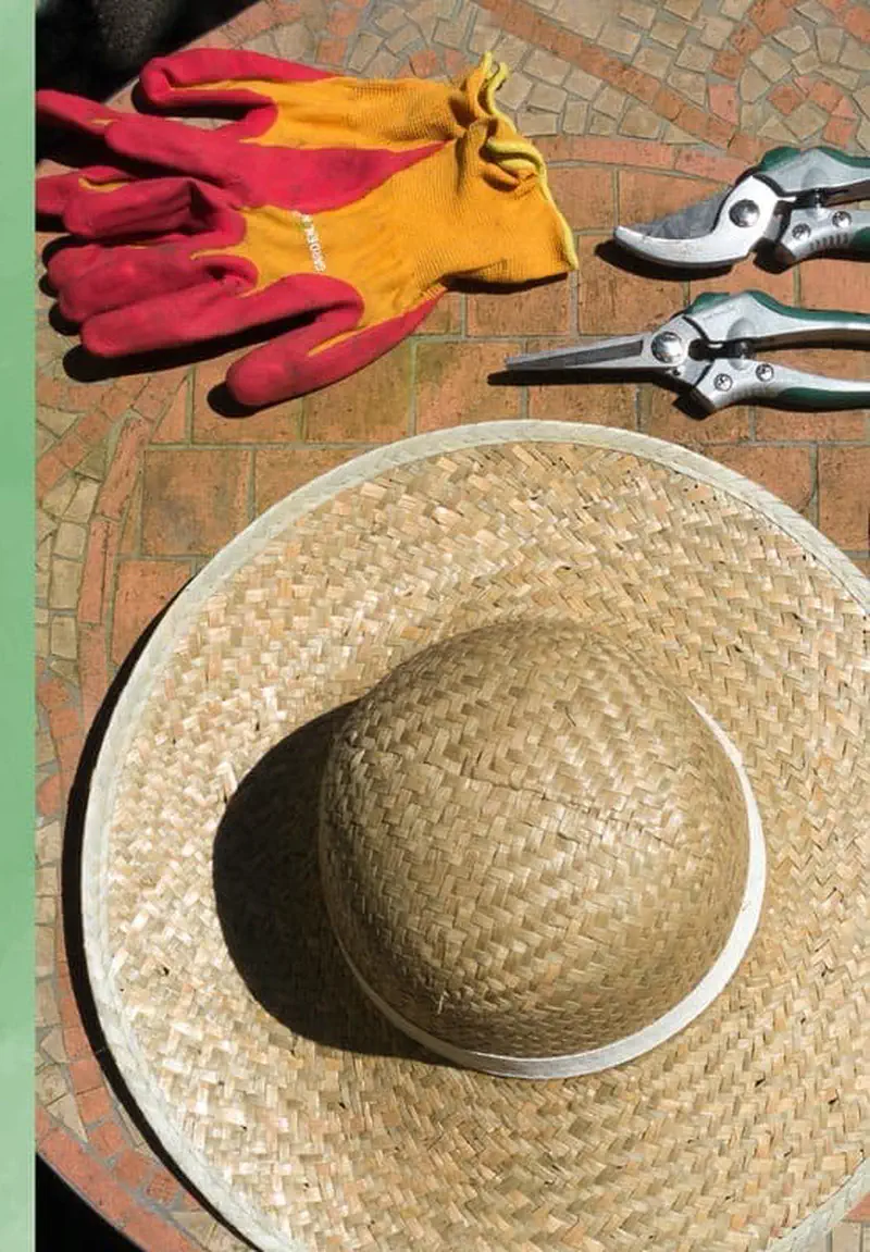 A wide-brimmed straw gardening hat on brick tiles next to orange and yellow gardening gloves and pruning shears.