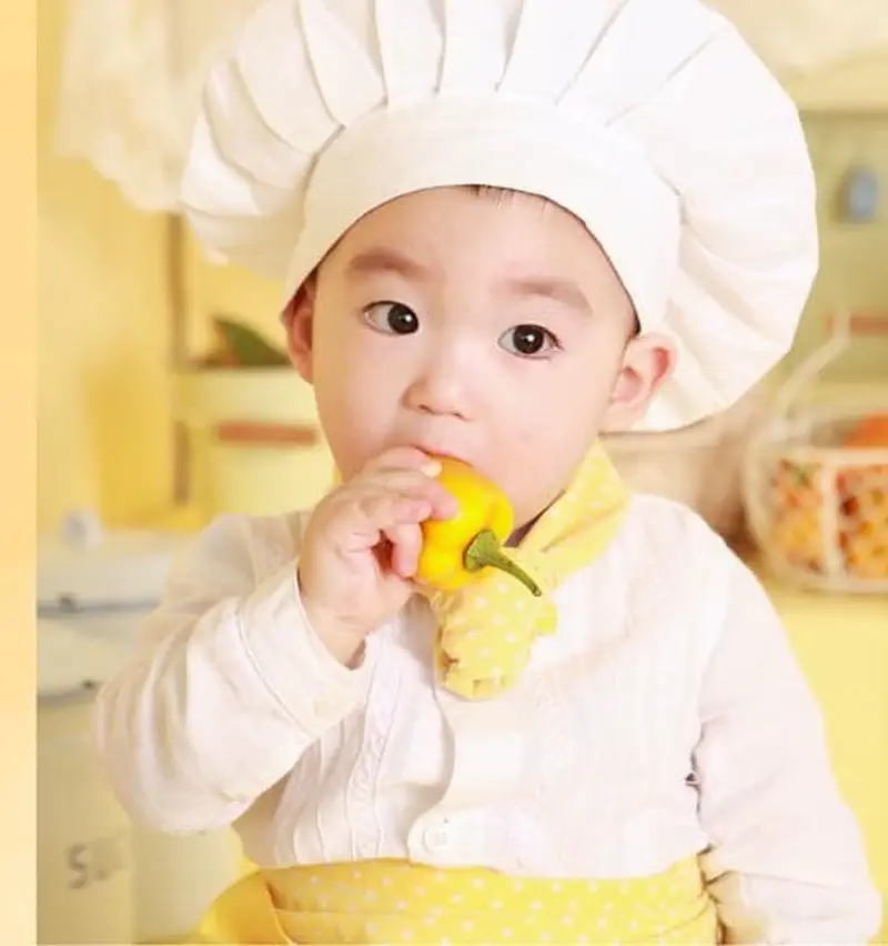 A baby wearing a white chef's hat and yellow apron, holding a yellow bell pepper up to their mouth in a kitchen.