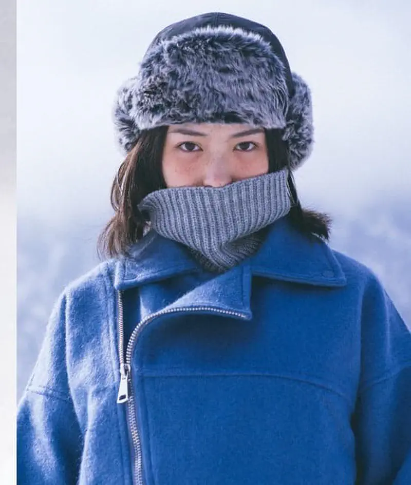 A woman wearing a grey furry winter hat with ear flaps and a grey knit scarf covering her face, in a blue coat.