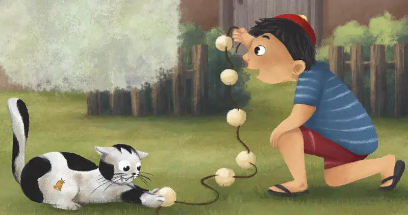Boy kneeling on grass playing with cat using string toy with pom poms by fence.
