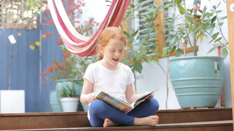 Child reading outdoors on a sunny porch, sounding out words from a picture book