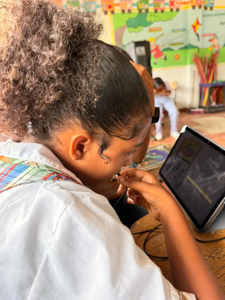 A student in Colombia holds the headset microphone near their mouth while reading a Spanish book aloud to Bookbot on a tablet, with a colorful alphabet mural on the classroom wall behind